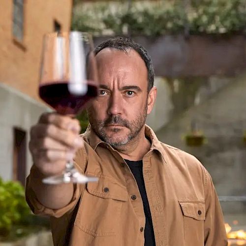 A man holds up a glass of red wine in an outdoor setting, focusing intently on it.