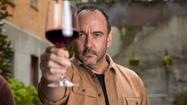 A man holds up a glass of red wine in an outdoor setting, focusing intently on it.