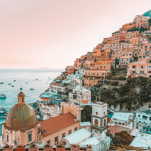 A picturesque coastal town with pastel-colored buildings on a hillside, overlooking a serene sea with boats under a warm sky.