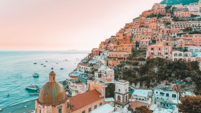A picturesque coastal town with pastel-colored buildings on a hillside, overlooking a serene sea with boats under a warm sky.