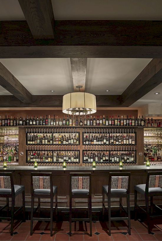 The image shows a well-stocked bar with high chairs and a warm, inviting interior featuring wooden beams and a chandelier.