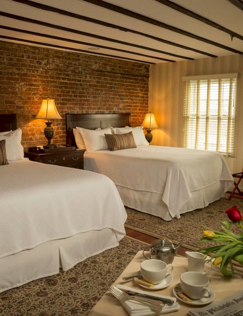 A cozy bedroom with two beds, bedside lamps, a brick wall, and a table set with breakfast items and flowers, creating a warm ambiance.