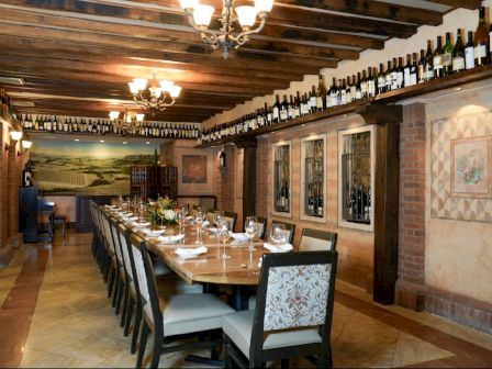 The image shows an elegant dining room with a long table set for a meal, surrounded by chairs and decorated with wine bottles and artwork.