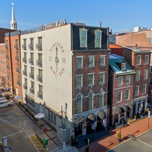 An aerial view of a brick-urban streetscape with a beige building featuring a clock, storefronts along a corner, and parked cars lining the curb, under a clear blue sky.
