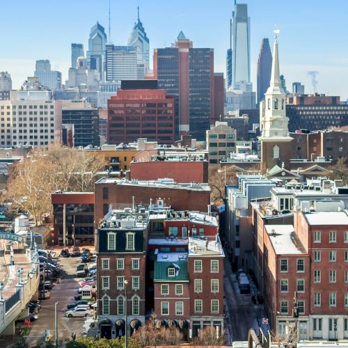 A city skyline with a mix of brick buildings in the foreground and modern high-rises in the background, under a clear blue sky.