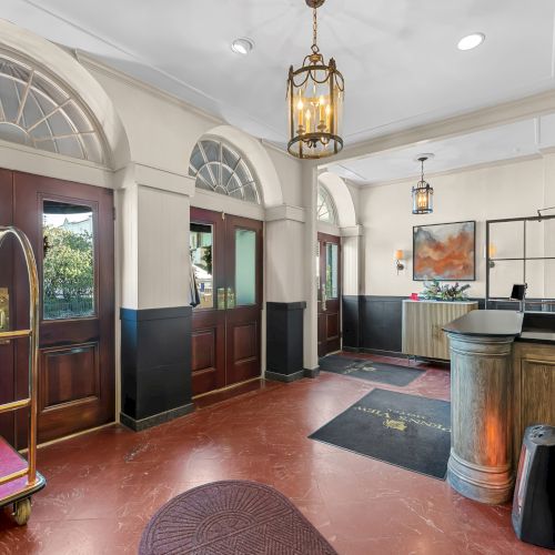 A hotel lobby with a front desk, a luggage cart, arched windows, wood doors, warm lighting, and plants, welcoming guests.