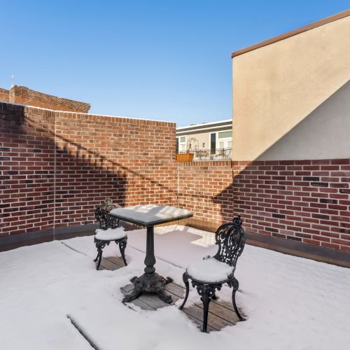A small rooftop terrace with a round table and two ornate metal chairs surrounded by brick walls, light snow on the floor, and clear blue sky.