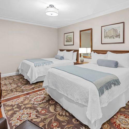 A cozy hotel room with two queen beds, wood headboards, a small desk, framed art, a dresser, and soft lighting.