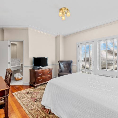 A bright, cozy hotel room with a large white bed, wooden furniture, a desk with a chair, a wall-mounted TV, and pastel walls; windows let in soft light.