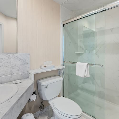 A modern bedroom with a white bed, glass shower enclosure, and neutral tones in a bright, minimalist bathroom suite.