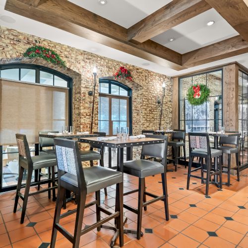 A cozy dining area with brick walls, arched windows, hanging wreaths, and wooden beams; high tables and chairs sit on orange tile floors, creating a rustic bistro vibe.