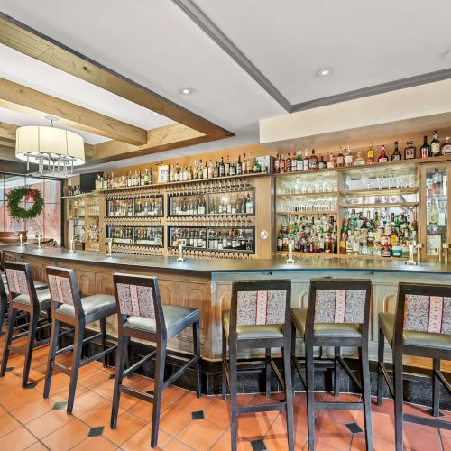 A stylish hotel bar with a long wooden counter, high stools, marble back bar, shelves stocked with bottles, warm lighting, and a wreath decor in the background.