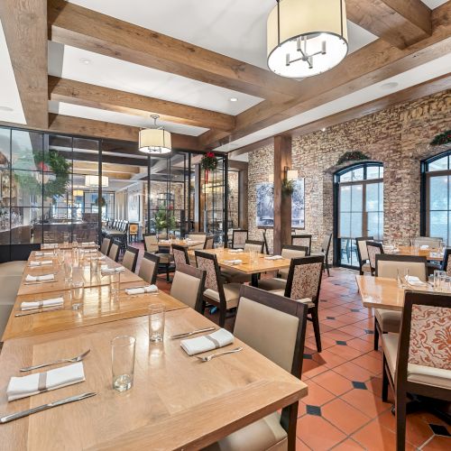 A stylish restaurant with wooden beams, brick walls, tiled floor, rowed tables set with cutlery, and large windows letting in light.