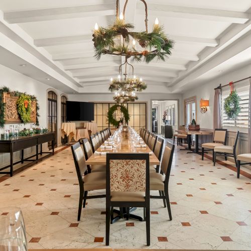 A long dining room with a central table, wooden chairs, festive greenery chandeliers, and wreaths along the walls, bright and elegant.