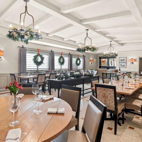 A bright, elegant restaurant with long wooden tables, upholstered chairs, wine glasses, napkins, and greenery decor in a white, airy room.