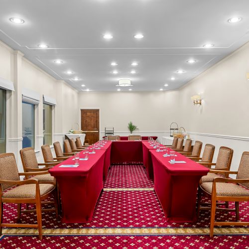 A long conference room with a U-shaped table setup, red tablecloths, beige chairs, white walls, and bright ceiling lights, ready for a meeting.