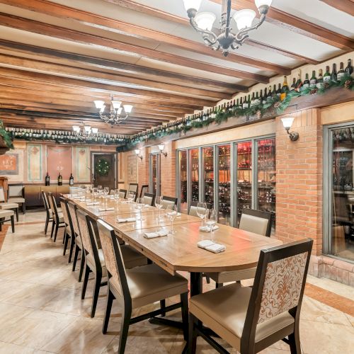 A long wooden dining table with matching chairs runs down a cozy, wine-cellar-style room lined with wine racks and glass-fronted cabinets.