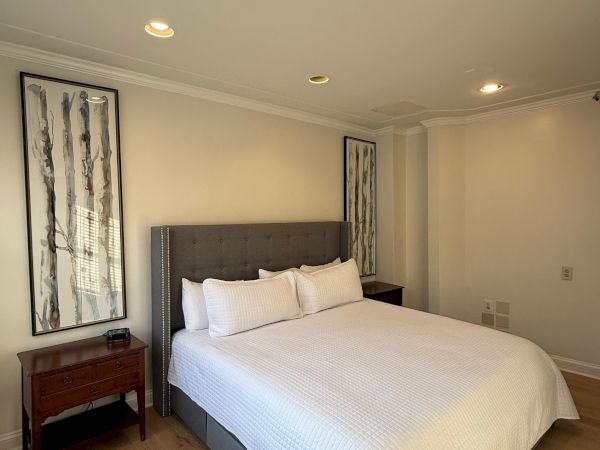 A neatly made bedroom with a gray upholstered bed, white bedding, wall art, and recessed ceiling lights.