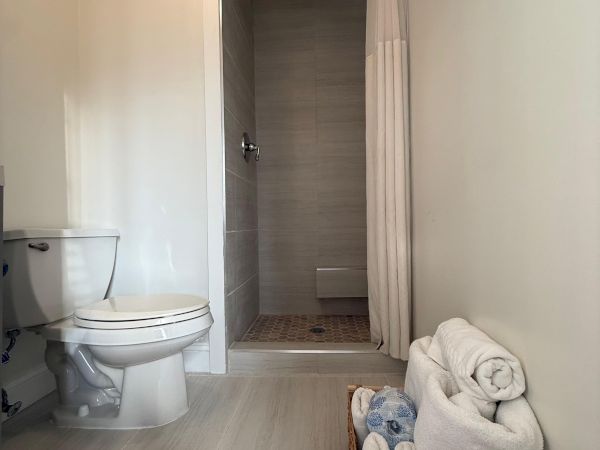 A bathroom with a toilet on the left and an open shower stall with a gray rainfall head, white curtain, and rolled towels.