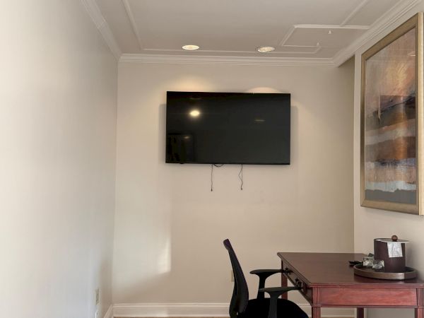 A bright room with recessed ceiling lights, a wall-mounted flat-screen TV, and a desk with a chair beneath.