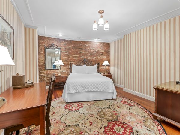 A styled bedroom with a white bed, striped wallpaper, exposed brick accent wall, bedside lamps, ceiling chandelier, and a patterned rug.