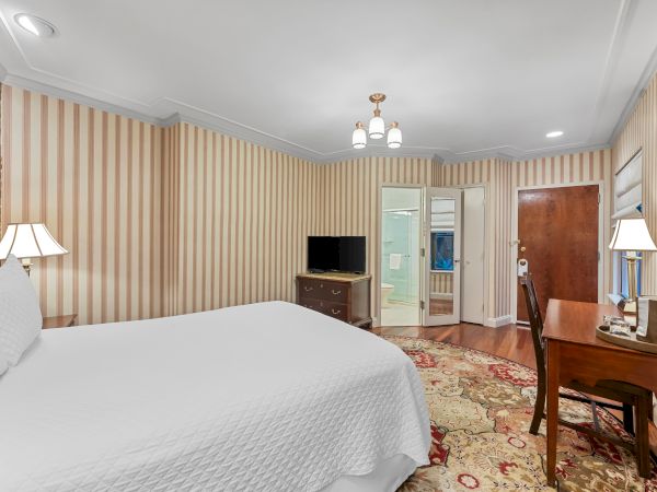 A cozy bedroom with striped walls, a bed, lamps, a dresser, TV, and a ceiling light, plus doors leading to a hallway.