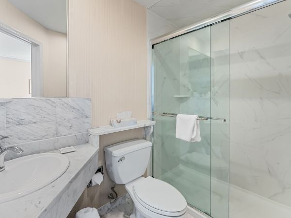 Bright modern bathroom with marble vanity, toilet, and a glass shower/tub with white towels and sleek lighting.