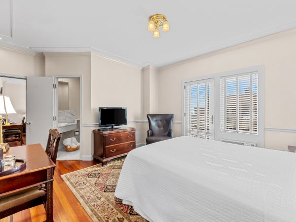 A bright bedroom with a large bed, TV on a dresser, warm ceiling light, windows with blinds, and an elegant rug.