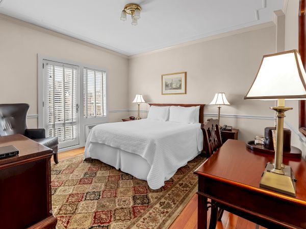 A bright bedroom with a large white bed, bedside lamps, framed wall art, a chandelier, and plantation shutters letting in sunlight.