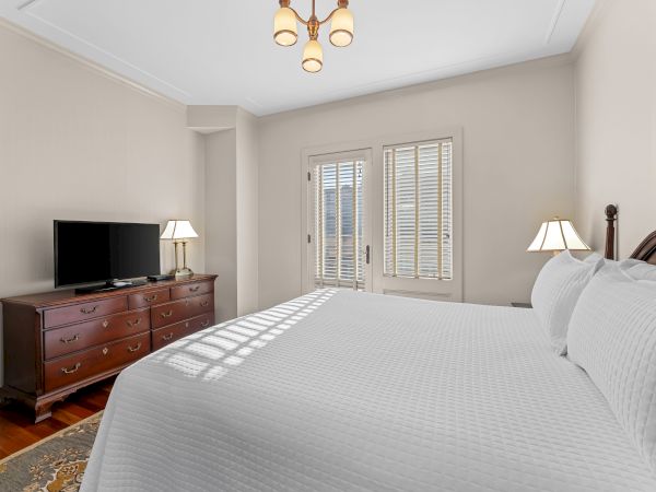 A bright bedroom with a large bed, TV on a dresser, bedside lamps, and a brass chandelier over white blinds.