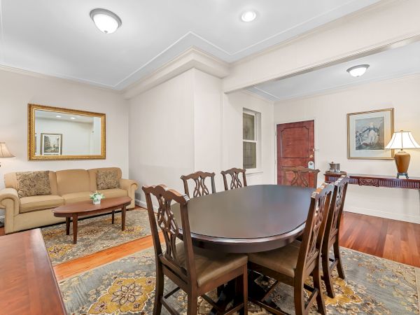 A bright, classic living/dining room with a round table, chairs, sofa, wall art, ceiling lights, and a wooden door.