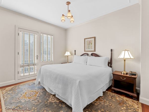 A bright bedroom with a white bed, bedside lamps, wood floor, patterned rug, and a ceiling chandelier above.