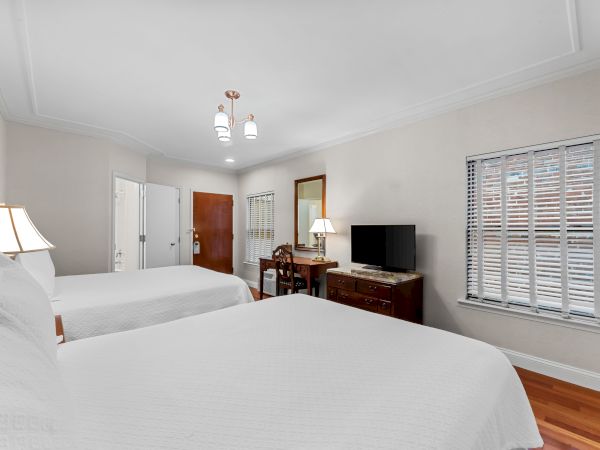A bright hotel bedroom with a bed, nightstands, a TV on a dresser, and large windows with white blinds.