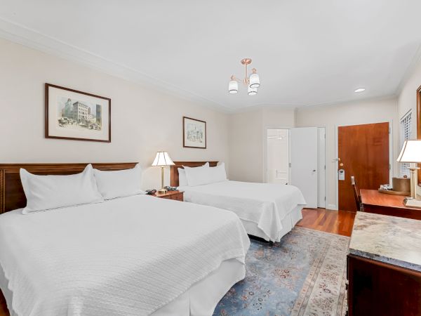 A bright hotel bedroom with two white beds, bedside lamps, wall art, a ceiling light fixture, and a wooden door and vanity area.