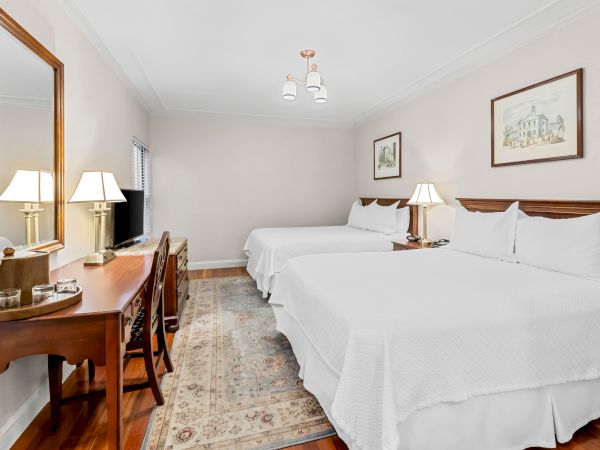 A bright hotel bedroom with two neatly made beds, a ceiling chandelier, lamps, framed wall art, and a wooden dresser.