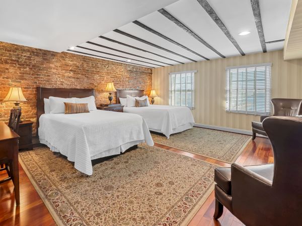 A bright hotel room with two twin beds, a brick accent wall, table lamps, large windows with blinds, and an armchair.