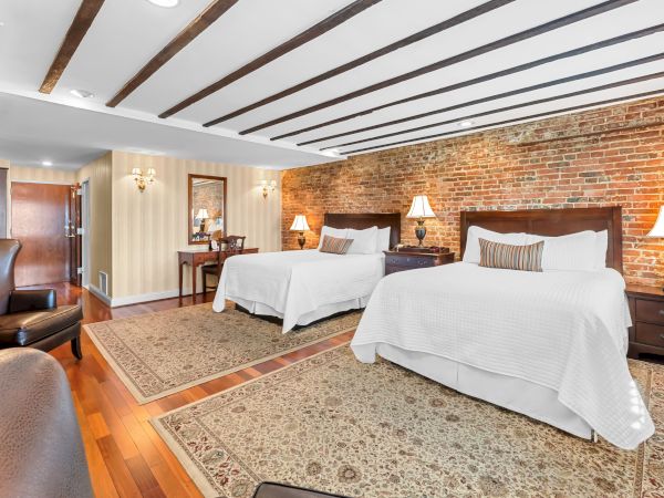 A cozy bedroom with two beds, white linens, wooden headboards, warm lamps, and exposed brick on the wall.