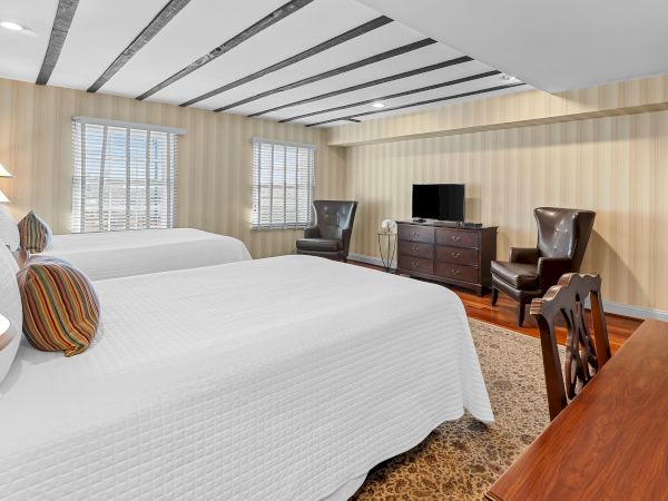 A cozy bedroom with two beds, wood slat walls, striped blinds, recessed ceiling lights, and a TV on a dresser beside leather chairs.