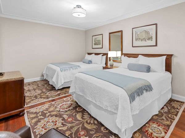 A bright hotel-style bedroom with two beds, blue pillows, a dresser, framed wall art, curtains, and a ceiling light.