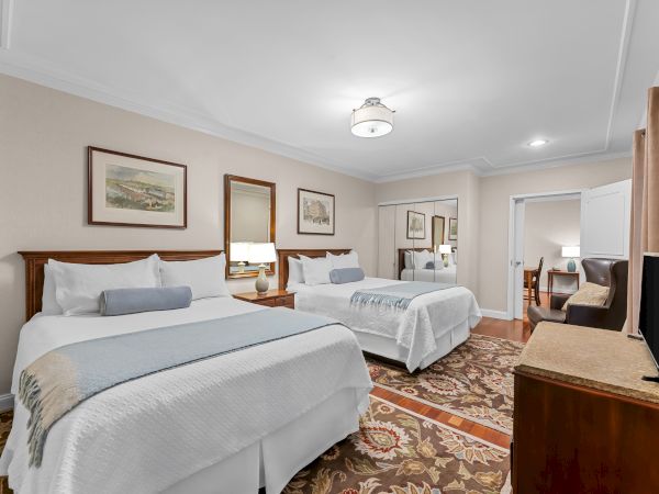 Bright hotel bedroom with two beds, bedside lamps, framed art, ceiling light, a curtain, and a desk with chair.