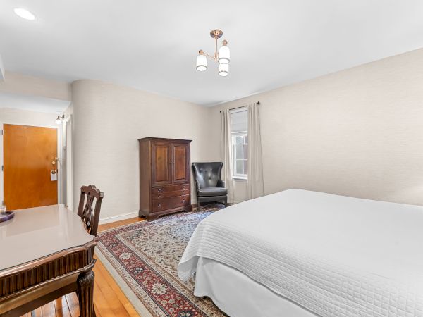 A bright bedroom with a bed, patterned rug, wooden armoire, chair, door, window, and a three-light ceiling chandelier.