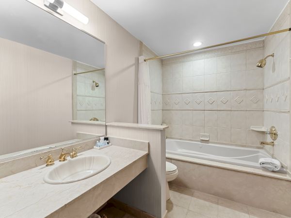 Bright bathroom with marble vanity, gold fixtures, tiled shower/tub, white curtain, ceiling light, and neatly folded towels.