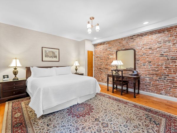 A cozy bedroom with a white bed, lamps, vintage desk and chair, and a red brick wall behind a large mirror.