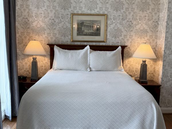 A classic hotel bedroom with a large white bed, two bedside lamps, ornate wallpaper, and a framed wall painting.