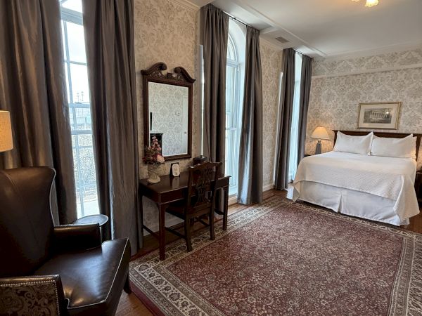 A classic hotel bedroom with patterned wallpaper, dark curtains, a king bed, a small desk, and a warm ceiling light.