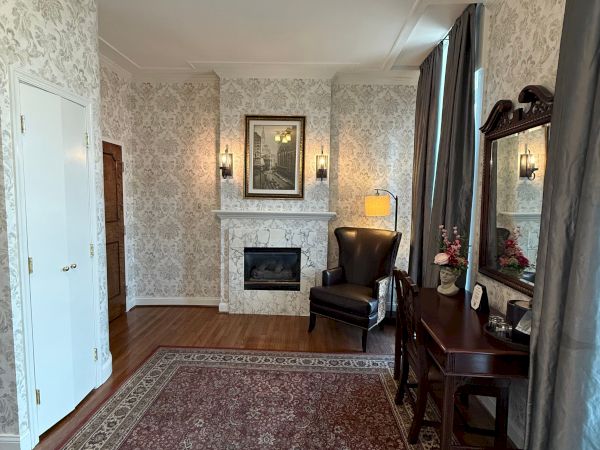 A cozy lounge with vintage wallpaper, a marble fireplace, framed art, a leather chair, curtained window, and an ornate rug.