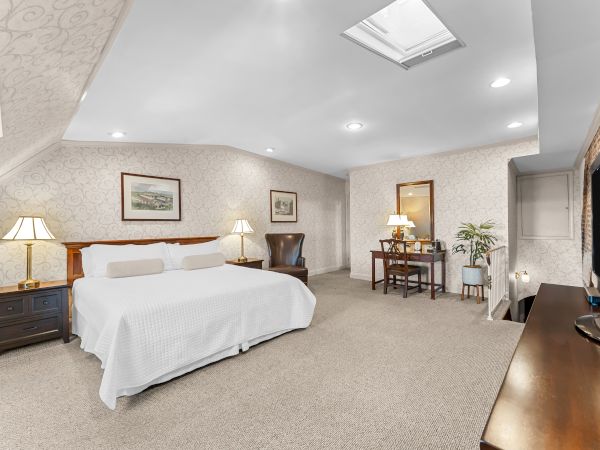 A bright hotel-style bedroom with a large bed, patterned wallpaper, bedside tables, chairs, desk, and ceiling lights.