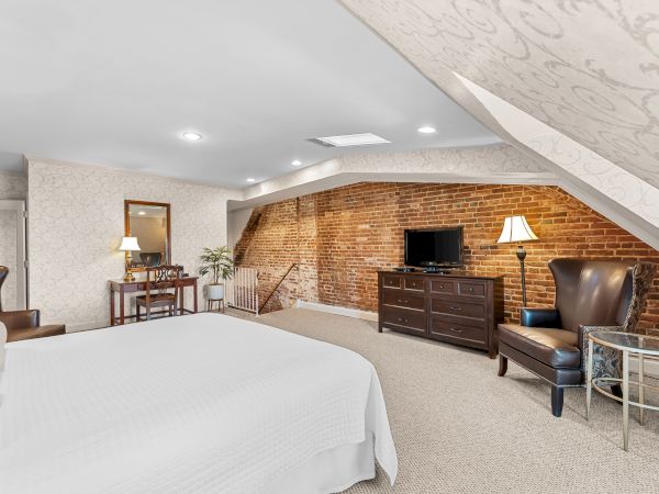 A cozy bedroom with a brick accent wall, flat-screen TV on a dresser, a bed, leather chairs, and warm lighting.
