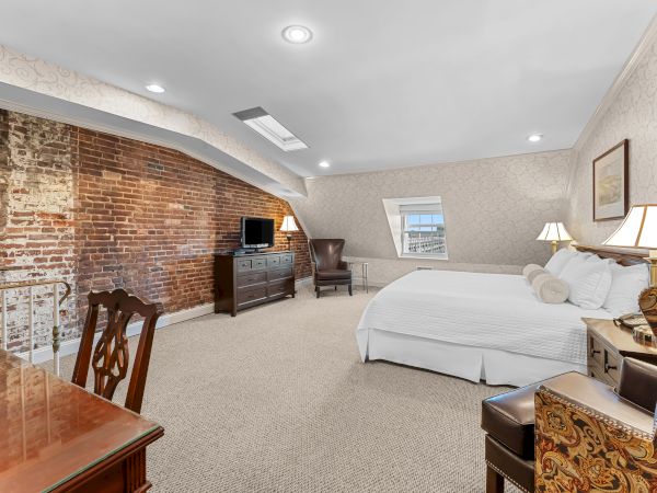 A stylish hotel bedroom with a large bed, lamps, a TV on a cabinet, exposed brick wall, and skylight beams under ceiling lights.