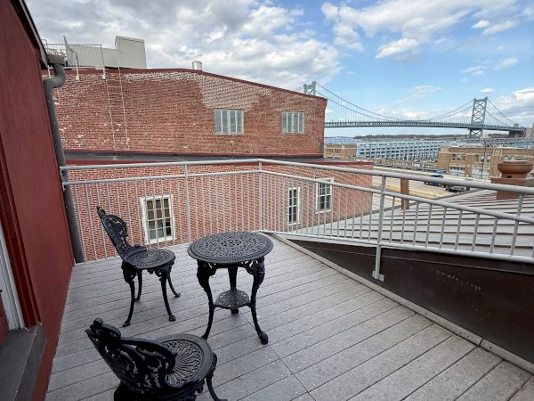 Rooftop patio with black chairs and a table, overlooking red brick buildings and a suspension bridge beneath cloudy skies.